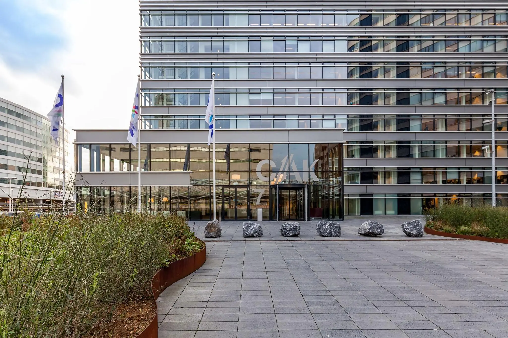 Vooraanzicht van het CAK-gebouw aan de Prinses Beatrixlaan met glazen entree, vlaggen en bestrate ingang met grote stenen.